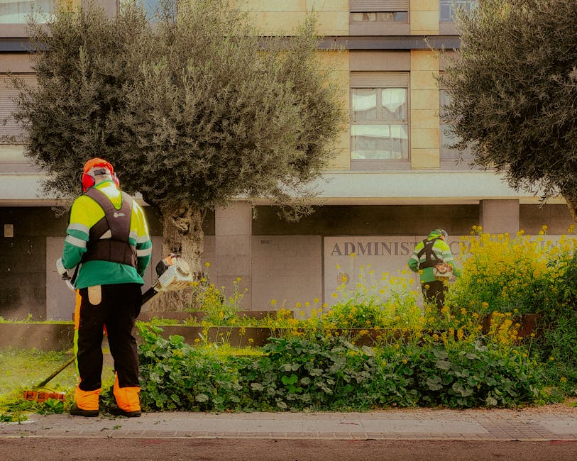 Two professional gardeners wearing high-visibility jackets and protective gear are working in a landscaped area outside a modern office building. One gardener, facing away from the camera, is using a 