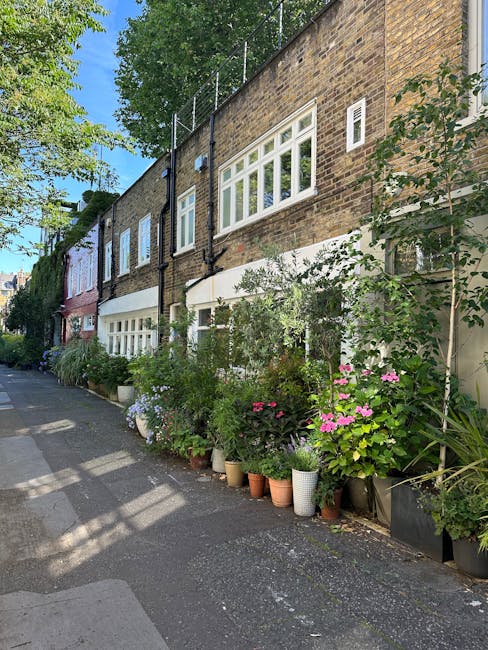 A well-maintained townhouse garden featuring a lush green lawn bordered by neatly trimmed hedges and flower beds with vibrant plants. In the foreground, there is a stone pathway leading to a small pat